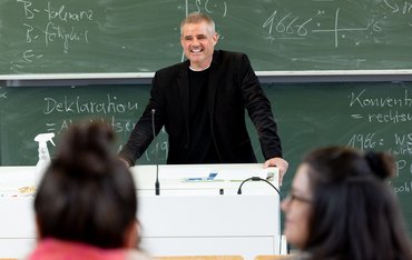 Ein Dozent steht an einem Rednerpult vor mehreren Studierenden.